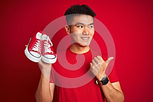 Young asian chinese man holding casual sneakers standing over isolated red background pointing and showing with thumb up to the