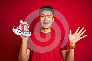 Young asian chinese man holding casual sneakers standing over isolated red background very happy and excited, winner expression