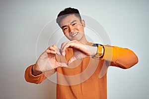 Young asian chinese man wearing orange sweater standing over isolated white background smiling in love showing heart symbol and
