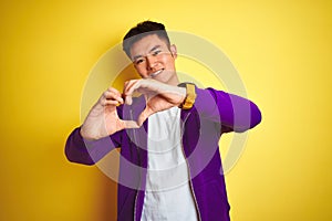 Young asian chinese man wearing purple sweatshirt standing over isolated yellow background smiling in love doing heart symbol