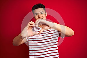 Young asian chinese man wearing striped t-shirt standing over isolated red background smiling in love doing heart symbol shape
