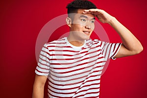 Young asian chinese man wearing striped t-shirt standing over isolated red background very happy and smiling looking far away with