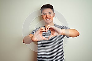 Young asian chinese man wearing striped t-shirt standing over isolated white background smiling in love showing heart symbol and