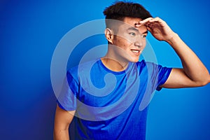 Young asian chinese man wearing t-shirt standing over isolated blue background very happy and smiling looking far away with hand