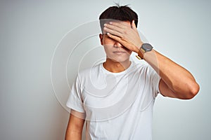 Young asian chinese man wearing t-shirt standing over isolated white background covering eyes with hand, looking serious and sad photo