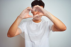 Young asian chinese man wearing t-shirt standing over isolated white background Doing heart shape with hand and fingers smiling