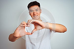 Young asian chinese man wearing t-shirt standing over isolated white background smiling in love doing heart symbol shape with