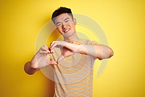 Young asian chinese man wearing t-shirt standing over isolated yellow background smiling in love doing heart symbol shape with