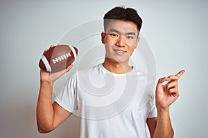 Young asian chinese sportsman holding rugby ball standing over isolated white background very happy pointing with hand and finger
