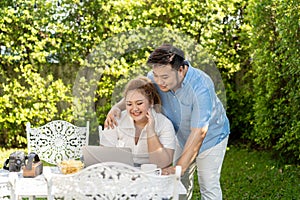 Young Asian chubby couple using a laptop in the outdoors green park together. Happy smiling man and woman working