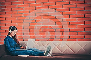 Young Asian college student girl using laptop computer, headphone, smartphone, notebook on couch sofa, sit at university campus