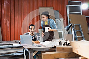 Young Asian couple carpenters man and woman discussing about design of products on laptop in woodcraft carpentry workshop.