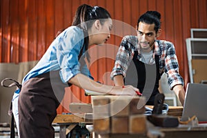 Young Asian couple carpenters man and woman discussing about design of products on laptop in woodcraft carpentry workshop.