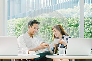 Young Asian couple, coworkers, or business partners have fun using smartphone together, with laptop computer at coffee shop