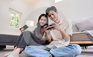 A young Asian couple happily sits together in the living room playing mobile games. Couple making romantic love in the