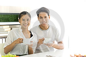 Young asian couple holding white coffee cup while look at camera smiling. Husband and wife sit at breakfast table.