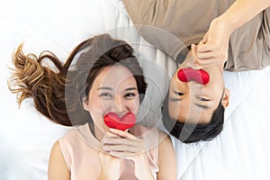 Young asian couple laying down in opposite direction together kissing red heart shape pillows on white sheet bed happily