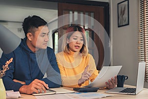 Young asian couple managing finances, reviewing their bank accounts using laptop computer and calculator at modern home.