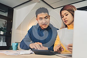 Young asian couple managing finances, reviewing their bank accounts using laptop computer and calculator at modern home.