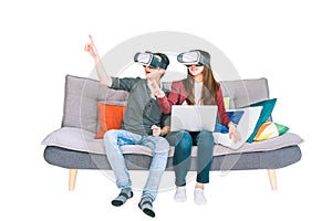 Young Asian couple playing VR virtual reality gadget, sitting on sofa together, isolated on white background. Modern gaming tech