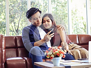 Young Asian couple sitting close together  on couch in living room looking at screen of mobile phone in a man`s hand