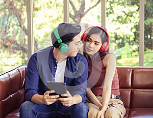 Young Asian couple sitting close together on couch ,looking happily to each other ,wearing headphones  listening to music from