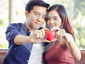 Young Asian couple sitting close together  holding red hearts in their hands. Selective focus on red heart