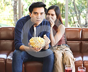 Young Asian couple sitting on couch in living room , watching funny comedy movie,a man holding popcorn in his hand while his