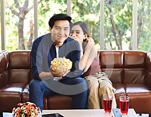 Young Asian couple sitting on couch in living room , watching funny comedy movie,a man holding popcorn in his hand while his