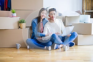 Young asian couple sitting on the floor of new house arround cardboard boxes using laptop and drinking a cup of coffee