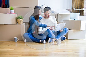 Young asian couple sitting on the floor of new house arround cardboard boxes using laptop and drinking a cup of coffee