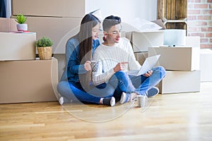 Young asian couple sitting on the floor of new house arround cardboard boxes using laptop and drinking a cup of coffee