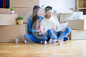 Young asian couple sitting on the floor of new house arround cardboard boxes using laptop and drinking a cup of coffee