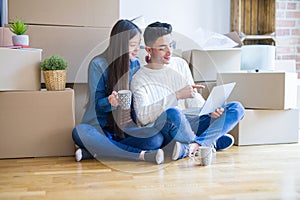 Young asian couple sitting on the floor of new house arround cardboard boxes using laptop and drinking a cup of coffee