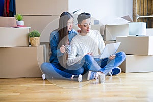 Young asian couple sitting on the floor of new house arround cardboard boxes using laptop and drinking a cup of coffee