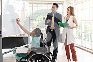 young asian disabled working woman sitting in wheelchair presenting her job with her colleagues in the working place, office room