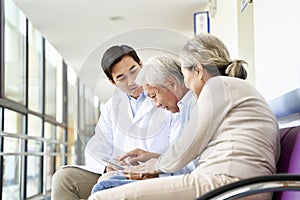 Young asian doctor talking to senior couple patients in hospital hallway