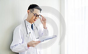 Young  asian doctor man wearing lab coat looking down reading to his patient notes standing in hospital