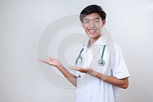 Young Asian doctor man wearing medical uniform, smiling cheerful presenting with palm of hand looking at the camera