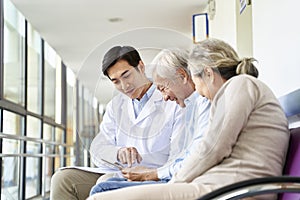 Young asian doctor talking to senior couple patients in hospital hallway