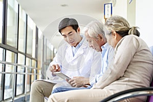 Young asian doctor talking to senior couple patients in hospital hallway