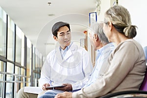 Young asian doctor talking to senior couple patients in hospital hallway