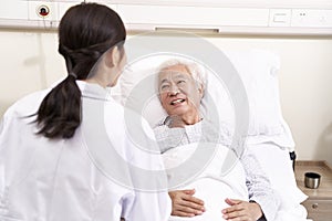 Asian old man lying in bed talking to young female doctor in hospital ward