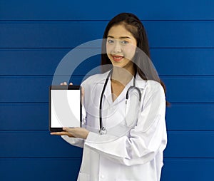 Young asian doctor in white gown and stethoscope holding white screen tablet computer, standing in front of blue wall