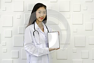 Young asian doctor in white gown and stethoscope holding white screen tablet computer, standing in front of white rectangle