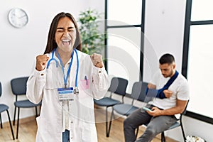 Young asian doctor woman at waiting room with a man with a broken arm excited for success with arms raised and eyes closed