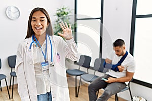 Young asian doctor woman at waiting room with a man with a broken arm showing and pointing up with fingers number five while