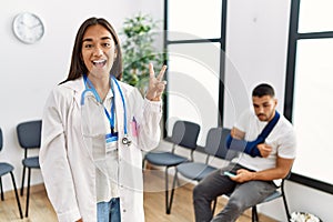 Young asian doctor woman at waiting room with a man with a broken arm showing and pointing up with fingers number two while
