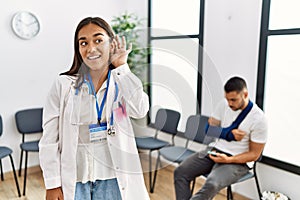 Young asian doctor woman at waiting room with a man with a broken arm smiling with hand over ear listening an hearing to rumor or