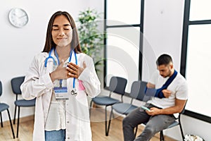 Young asian doctor woman at waiting room with a man with a broken arm smiling with hands on chest with closed eyes and grateful